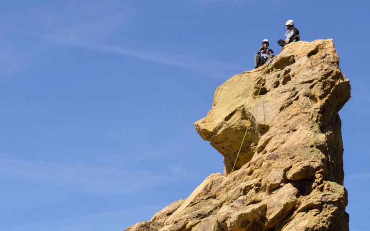 two people wearing rock climbing gear sit high atop a tall rock wall after climbing it